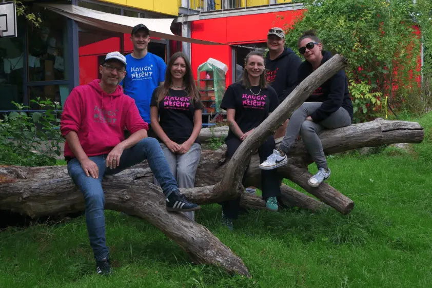 Das sechsköpfige Team von Mauga posiert auf einem großen Baumstamm im Freien. Sie tragen größtenteils Kleidung mit dem Magenta-Schriftzug „MAUGA NESCHT“.