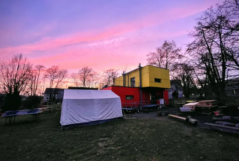 Ein großes weißes Zelt steht auf einer Wiese vor einem markanten Gebäude mit rotem Erdgeschoss und gelbem Obergeschoss. Im Hintergrund leuchtet der Abendhimmel in intensiven Pink- und Violetttönen.