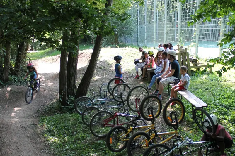 Kinder sitzen an der BMX-Bahn auf Bänken im Grünen und schauen den vorbeifahrenden Rasfahrern zu. Vor ihnen stehen einige Fahrräder fahrbereit.