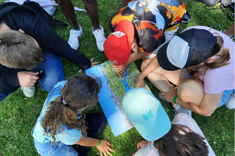 Eine Gruppe von Kindern hockt auf einer Wiese im Kreis um eine ausgebreitete Landkarte und betrachtet sie aufmerksam. Ein Kind zeigt mit dem Finger auf einen bestimmten Punkt.