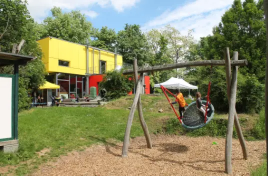 Zwei Kinder schaukeln auf einer Nestschaukel auf einem Spielplatz vor einem modernen gelb-roten Gebäude im Grünen.