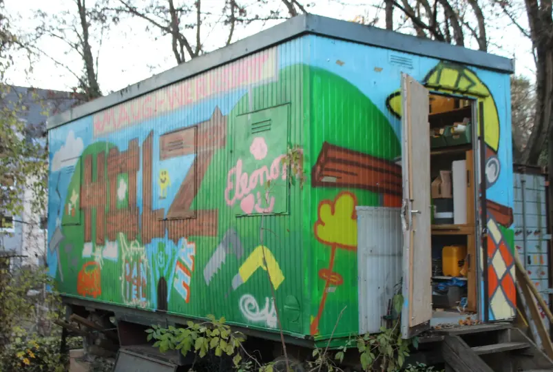 Ein alter Waggon, das mit Graffiti bunt bemalt ist und als Holz-Werksatt dient. Die Tür des Waggons steht halb offen und im Innern ist ein Regal mit Materialien und Werkzeugkoffern zu sehen.
