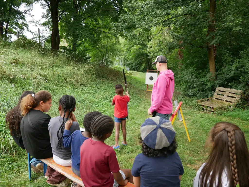 Auf einer Holzbank sitzende Kinder beobachten, wie ein Kind unter Anleitung eines Erwachsenen mit Pfeil und Bogen auf eine Zielscheibe schießt.