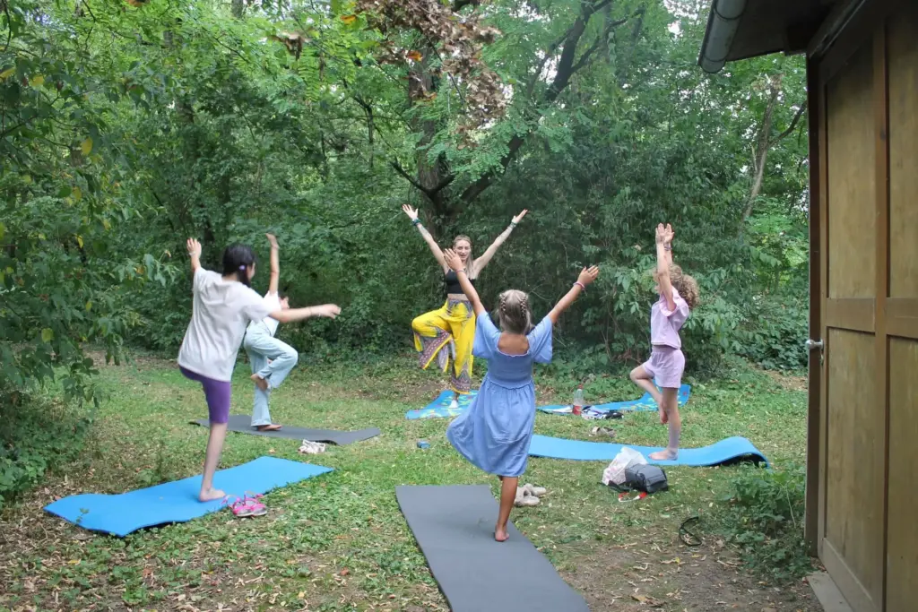 Eine Yogagrupe, bestehend aus einer Yogalehrerin und vier Mädchen, übt die Baumpose im Freien auf Matten, umgeben von dichtem Grün.