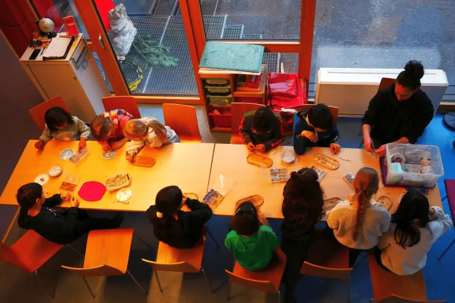 Aus der Vogelperspektive zu sehen ist eine Gruppe Kinder, die um zwei Tische sitzt und an Kunst- und Bastelprojekten arbeitet, wobei eine erwachsene Person am Tisch mitsitzt und ihnen hilft.