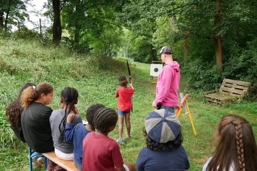 Auf einer Holzbank sitzende Kinder beobachten, wie ein Kind unter Anleitung eines Erwachsenen mit Pfeil und Bogen auf eine Zielscheibe schießt.
